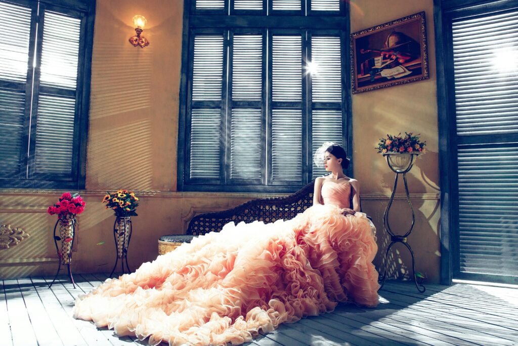 A bride in a luxurious gown sits gracefully in a sunlit room, exuding elegance.