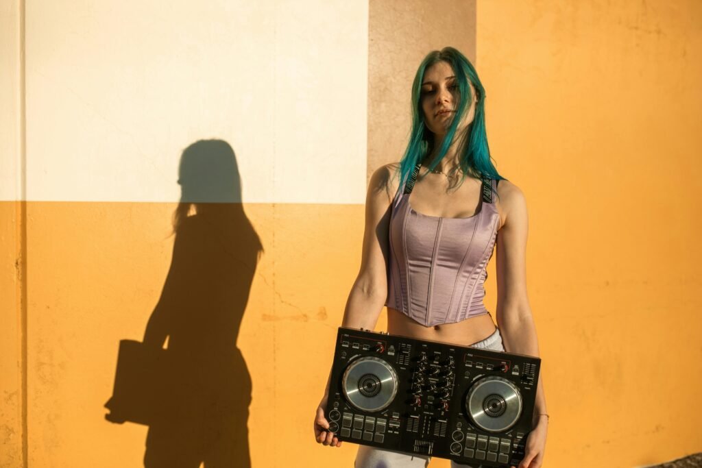 Young DJ with blue hair holding a mixer, posing stylishly in urban daylight.