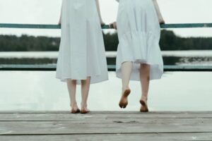 Two women in white dresses stand barefoot on a lakeside dock, enjoying a serene moment.