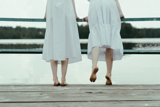 Two women in white dresses stand barefoot on a lakeside dock, enjoying a serene moment.